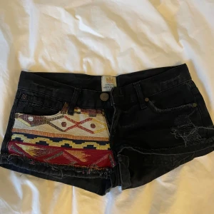 Svarta jeansshorts med broderad detalj från River Island - Unika svarta jeansshorts från River Island med färgglad broderad panel framtill. Klassisk femficksmodell, lågmidjade och råa kanter. Perfekta för sommaren när du vill sticka ut lite extra.