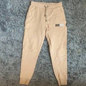 Beige mjukisbyxor från Hoodrich - Säljer ett par beige mjukisbyxor från Hoodrich i storlek medium. Byxorna har resår och snörning i midjan, muddar vid bensluten och en tryckt logga på ena benet. Perfekta för en avslappnad stil.