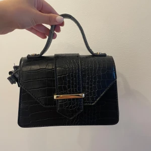 Svart handväska  - Snygg svart handväska med krokodilpräglat mönster och guldfärgad detalj på locket. 