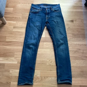 Blå jeans från Nudie Jeans - Säljer ett par klassiska blå jeans från Nudie Jeans med orangea sömmar och snygga bakfickor med vågdetalj. Modellen är Grim Tim med normal passform och raka ben. Materialet är 99% bomull och 1% elastan för lite stretch.
