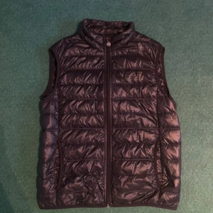 Svart dunväst från Emporio Armani EA7 - Säljer en svart dunväst från Emporio Armani EA7 med hög krage och dragkedja framtill. Västen har två sidofickor och diskret logga på bröstet. Perfekt för lager-på-lager och stilren look. Inga defekter mycket fin väst!