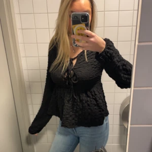Svart långärmad blus med knyt - Säljer en svart långärmad blus med knyt framtill. Blusen har en strukturerad, lite skrynklig yta och v-ringning. Storlek M men sitter bra på mig som har storlek S