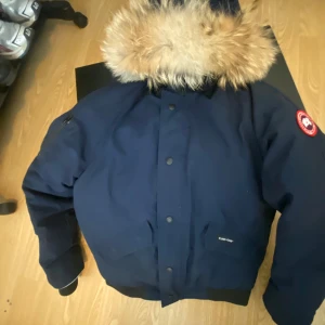 Mörkblå dunjacka från Canada Goose med äkta pälskrage - Säljer en mörkblå Canada Goose dunjacka med stor äkta pälskantad huva och klassisk logga på ärmen. Jackan har knappar och dragkedja framtill samt fickor med lock och dragkedja. Perfekt för kalla dagar. Original låda och kvitto finns.