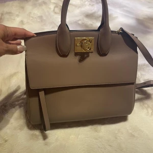 Beige Ferragamo Studio Bag - Säljer en lyxig beige handväska från Ferragamo med guldfärgat spänne och två handtag. Väskan har en stilren design med lock och dekorativa metalldetaljer undertill. Perfekt för dig som vill ha en elegant accessoar.