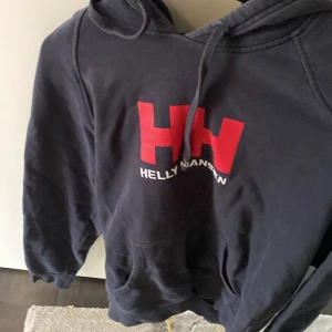 Mörkblå hoodie från Helly Hansen - Säljer en mörkblå hoodie från Helly Hansen med stor röd HH-logga och vit text på bröstet. Tröjan har huva med dragsko och en stor magficka. Perfekt för dig som gillar klassisk streetstil. 