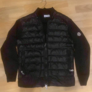 Svart Moncler cardigan - Säljer en svart pufferjacka från Moncler med quiltad framsida och ärmar i stickad bomull. Jackan har dragkedja framtill, två fickor med dragkedja och Moncler-logga på ärmen. Perfekt för våren och sena sommar kvällar. Jackan är i jätte bra skick. Pris kan diskuteras vid snabb affär. Fraktar inom 1 dag.