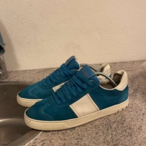 Blå och vita sneakers från Adidas - Säljer ett par blå sneakers från Adidas med vita detaljer och vit sula. Skorna har klassisk snörning och är tillverkade i mocka och syntet. Perfekta för dig som gillar en sportig och stilren look.