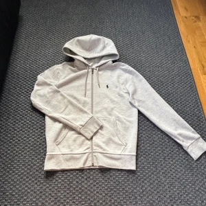Ljusgrå hoodie från Polo Ralph Lauren - Säljer en stilren ljusgrå hoodie från Polo Ralph Lauren med dragkedja och huva. Tröjan har två fickor framtill och en broderad logga på bröstet. Perfekt för en avslappnad look. Priset är inte hugget i sten!