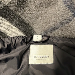  pufferjacka/väst  burberry - Säljer en svart pufferjacka från Burberry med stor logotyp på ärmen och klassiskt rutigt foder i huvan. Jackan har dragkedja och flera fickor framtill. Perfekt för kyliga dagar och riktigt snygg streetstil.