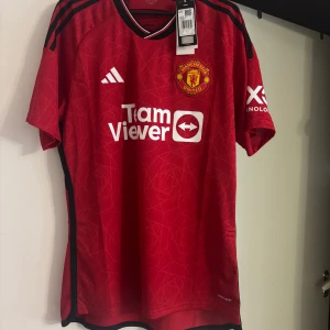 Manchester United hemmatröja 2023/24 från Adidas - Röd Manchester United hemmatröja från Adidas med klubbmärke på bröstet, svarta detaljer och vita logotyper. Kortärmad modell med rund hals och diskret mönster i tyget. Perfekt för fans som vill visa sitt stöd på matchdagar.