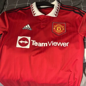 Manchester United hemmatröja Antony 21 - Röd Manchester United hemmatröja från Adidas med vitt krage och svarta detaljer. Tryck med TeamViewer, klubbmärke och Adidas-logga på framsidan. På ryggen står det Antony och nummer 21. Klassisk fotbollströja för fans.