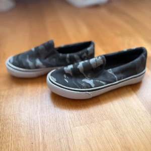 Svarta slip-on sneakers med camouflagemönster (barnskor) - Säljer ett par svarta slip-on sneakers med grått camouflagemönster och vit sula. Skorna har en rund tå och är perfekta för en avslappnad stil. Lätta att ta på och av tack vare elastiska paneler på sidorna.