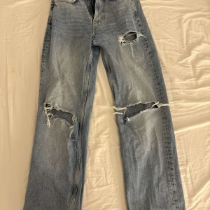 Ljusblå slitna jeans med raka ben - Säljer ett par ljusblå jeans med flera stora slitningar och hål på framsidan. Jeansen har raka ben och klassisk femficksdesign. Perfekta för dig som gillar en avslappnad och trendig streetstil.