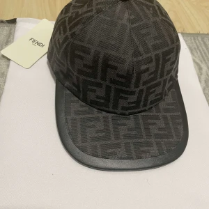 Svart keps från Fendi med monogram - Snygg svart keps från Fendi med det klassiska FF-monogrammet över hela kepsen. Kepsen har böjd skärm i svart och justerbar rem baktill med Fendi-logga i metall. Perfekt accessoar för en trendig look.