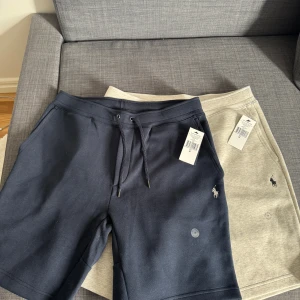 Shorts från Polo Ralph Lauren - Säljer två par shorts från Polo Ralph Lauren. Ett par är mörkblå och det andra är gråa. Båda har snörning i midjan och en liten broderad logga på framsidan. Perfekta för en avslappnad stil. ”Styck pris 1049”