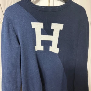 Blå stickad tröja från Tommy Hilfiger - Säljer en blå stickad tröja från Tommy Hilfiger med en stor vit bokstav H på bröstet. Tröjan har rund halsringning och långa ärmar. Perfekt för dig som gillar klassisk och stilren design.
