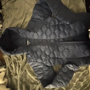 Mörkblå pufferjacka från STRÖVA - Mörkblå pufferjacka från STRÖVA i storlek XL. Jackan har huva, lång ärm och stängs med dragkedja. Quiltad design som ger en snygg och modern look. Perfekt för kyliga dagar. Köpt från Ullared för 299kr aldrig använt den innan 