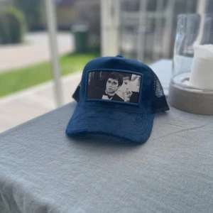 Blå keps med nät och tryck - Säljer en blå keps från Champone med svart nät baktill och justerbar snapback. Framtill finns ett svartvitt tryck med motiv. Kepsen har en klassisk böjd skärm och är perfekt för en avslappnad stil.