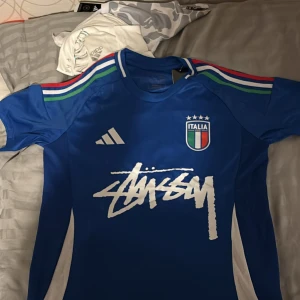Blå Italien fotbollströja Adidas x Stüssy - Säljer en blå Italien fotbollströja från Adidas i samarbete med Stüssy. Tröjan har korta ärmar, Italiens klubbmärke på bröstet, Adidas-logga och Stüssy-tryck framtill. Ränder i grönt, vitt och rött på axlarna. Helt ny 
