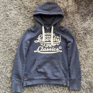 Blå hoodie från Superdry - Säljer en blå hoodie från Superdry med vit tryckt logga på bröstet och klassisk känguruficka. Tröjan har huva med vita snören och mjukt fleecefoder på insidan. Perfekt för en avslappnad stil.