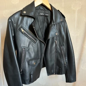 Svart skinnjacka från Zara - Snygg svart skinnjacka från Zara med klassisk bikerstil. Jackan har flera silverfärgade dragkedjor, bred krage och tryckknappar. Jackan är figursydd och har långa ärmar. Använd några gånger, saknar bälte och har inga andra fel. 