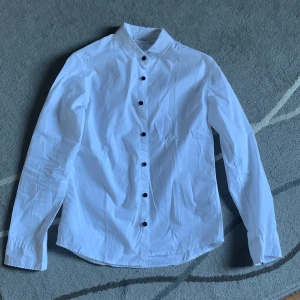 Vit Kappahl skjorta med svarta knappar - Klassisk vit skjorta med svarta knappar och lång ärm. Skjortan har en rak passform och är tillverkad i bomull, perfekt för en clean och stilren look. Passar till många olika outfits och är enkel att matcha.