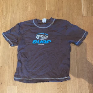 Brun t-shirt med blått tryck - Brun t-shirt/baby tee med kontrastsömmar och blått och beiget tryck framtill. Egentligen en barn tröja men den sitter jätte snyggt på