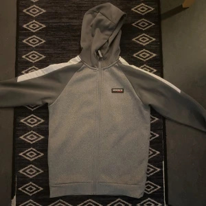 Grå hoodie med dragkedja från McKenzie - Säljer en grå hoodie från McKenzie med hel dragkedja och huva. Jackan har vita detaljer längs axlarna och ärmarna. Perfekt för en sportig stil och enkel att matcha med det mesta.