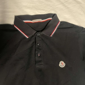 Svart pikétröja från Moncler - Säljer en svart pikétröja från Moncler med klassisk krage och knäppning framtill. Tröjan har Moncler-logga på bröstet och detaljer i rött och vitt på kragen. 