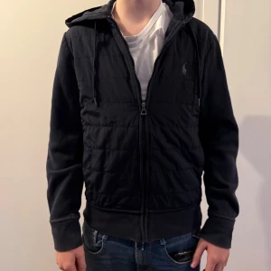 Blå cardigan från Polo Ralph Lauren - Mörkblå cardigan från Polo Ralph Lauren med dragkedja och huva. Jackan har ribbade muddar och en diskret logga på bröstet. 