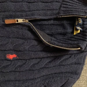 Mörkblå stickad tröja med dragkedja från Polo Ralph Lauren - Säljer en mörkblå kabelstickad tröja från Polo Ralph Lauren med halv dragkedja och röd broderad logga på bröstet. Den är lite liten i strolek, men är äkta. Skicka om det skulle vara några funderingar
