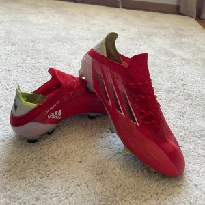 Röda Adidas fotbollsskor - Säljer ett par röda adidas X SPEEDFLOW.1  med vita och silverfärgade detaljer samt svarta ränder på sidorna. Skorna har snörning och är designade för att ge bra grepp på planen. Perfekta för dig som vill sticka ut på fotbollsplanen! Skick 9/10 Hör av dig för mer frågor och funderingar!