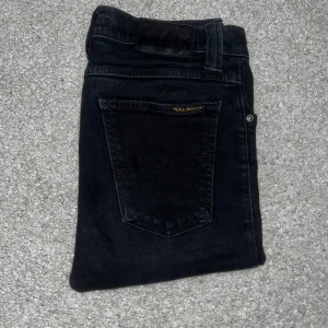Svarta slim fit jeans från nudie - Säljer ett par svarta skinny jeans från Replay med klassisk femficksdesign och diskret logga på bakfickan. Jeansen har normal passform i midjan och slim hela vägen ner. Perfekta för en stilren look. 28/32