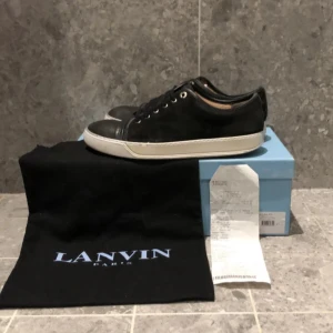 Svarta sneakers från Lanvin - Svarta sneakers från Lanvin med vit platt sula och rund tå. Skorna har snörning och metallfärgade öljetter. Klassisk och stilren design med diskret logga på sidan. Kommer med originalkartong och dustbag. Priset går att gå ner vid snabb affär