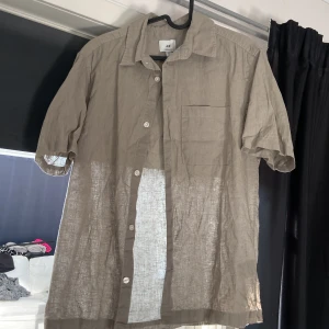 Grön linneskjorta med kort ärm - Säljer en luftig beige skjorta i linne från H&M. Skjortan har korta ärmar, klassisk krage, bröstficka och knäppning framtill. Perfekt för varma dagar när du vill ha en avslappnad men stilren look.