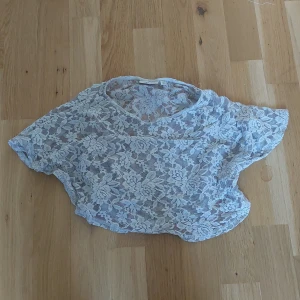 Vit spetsblus med blommönster - Säljer en söt vit blus i spets med blommigt mönster. Blusen är kortärmad och har en rund halsringning. Perfekt att styla över ett linne eller bh för en trendig look.