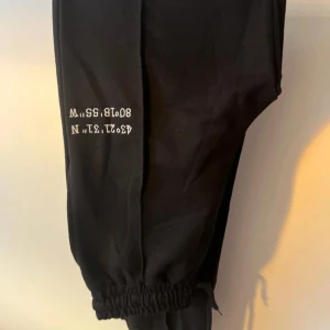 Svarta mjukisbyxor med tryck - Svarta mjukisbyxor med resår vid benslut och vitt tryck med siffror och bokstäver på ena benet. Perfekta för en avslappnad stil och riktigt sköna att ha på sig.