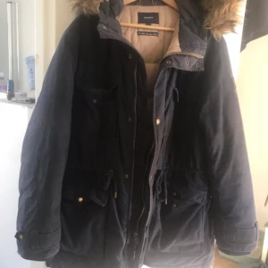 Mörkblå parkajacka med päls från Gant - Snygg mörkblå parkajacka från Gant med huva och päls. Jackan har dragkedja, knappar och flera fickor framtill. Perfekt för kyliga dagar och har en klassisk look med justerbar midja.