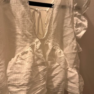 Vit volangblus med knytning - Säljer en söt vit blus med volanger och djup v-ringning. Blusen har knytning i ryggen och är tillverkad i ett lätt, krinklat tyg. Perfekt för varma dagar och ger en romantisk känsla till outfiten.