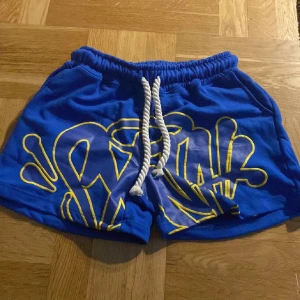 Blåa shorts med gul grafisk design och snörning - Snygga blå shorts med en stor gul grafisk print framtill och vit snörning i midjan. Perfekta för sommardagar eller strandhäng. Resår i midjan för skön passform.