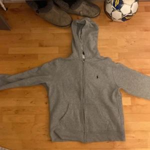 Grå hoodie från Polo Ralph Lauren - Säljer en klassisk grå hoodie från Polo Ralph Lauren med dragkedja och huva. Tröjan har två fickor framtill och den ikoniska broderade loggan på bröstet. Perfekt för en avslappnad stil.