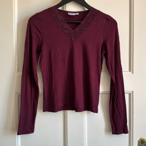 Vinröd långärmad topp med spets från Gina Tricot - Säljer en vinröd långärmad topp från Gina Tricot med v-ringning och fina spetsdetaljer vid halsen. Perfekt att styla till jeans eller kjol för en snygg look.