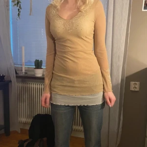 Beige långärmad topp med spetsdetaljer - Säljer en beige långärmad topp i mesh material med v-ringning och fina spetsdetaljer samt broderat mönster framtill. Tunn och stretchig modell som passar från S till L beroende på hur man vill att den ska sitta!