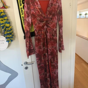 Blommig långklänning med slits och v-ringning - Säljer en långklänning i rödrosa toner med blommigt mönster. Klänningen har v-ringning, långa ärmar och en hög slits framtill. Snygg omlottkänsla vid midjan och lätt utsvängda ärmslut. Perfekt för speciella tillfällen!