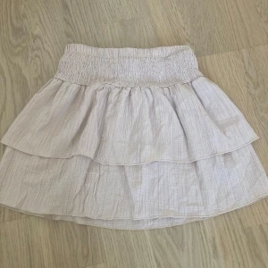 Beige volangkjol med resårmidja - Beige volangkjol med resårmidja. Söt beige/ljusrosa kortkjol med volanger och smockad resår i midjan. Perfekt för varma dagar. Lätt och luftig modell som ger en avslappnad och trendig look.