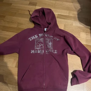 Vinröd hoodie från One of One - Säljer en vinröd hoodie med dragkedja från One of One. Tryck på bröstet med texten 'The World of Moominvalley' och motiv. Tröjan har huva och två fickor framtill. Perfekt för en avslappnad stil.