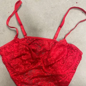 Röd spetsig korsettopp - Säljer en röd korsettopp med spetsdetaljer och justerbara axelband. Toppen har en figurnära passform och är perfekt för att lyfta din outfit med en färgstark touch. Spetsmaterialet ger en romantisk och trendig känsla.
