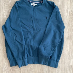 Blå sweatshirt från U.S. Polo Assn. - Säljer en stilren blå sweatshirt från U.S. Polo Assn. med rund halsringning och broderad logga på bröstet. Tröjan har långa ärmar och ribbade muddar vid ärmslut och nederkant. Perfekt för en avslappnad look.