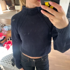 Mörkblå croppad stickad tröja - Säljer en mörkblå croppad stickad tröja med hög hals och långa ärmar. Tröjan har ribbade muddar vid ärmslut och nederkant samt ett snyggt stickat mönster längs axlarna. 