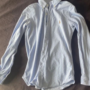 Blå- och vitrandig skjorta från Ralph Lauren - Klassisk skjorta med blå och vita ränder från Ralph Lauren. Skjortan har lång ärm, knappar framtill och en broderad logga på bröstet. Perfekt för en stilren och tidlös look.